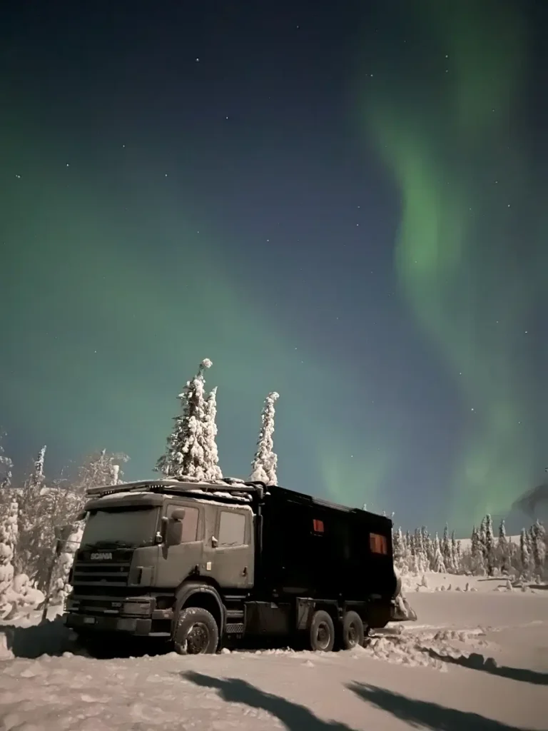 Geländegängiges Expeditionsmobil im Schnee bei Nacht unter den Nordlichtern.