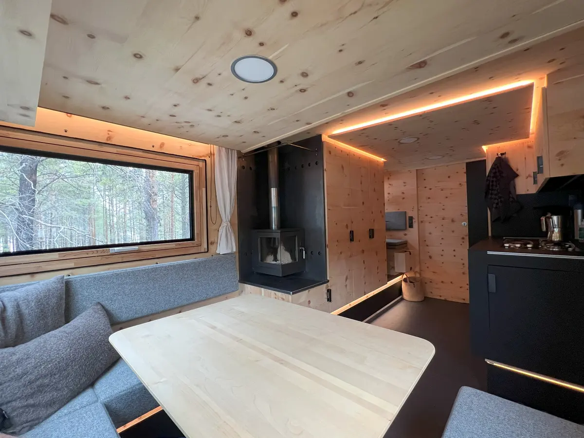 Der Innenraum eines stilvollen Selbstausbau-Campervans mit Holzkamin, Sitzgruppe und Waldblick.