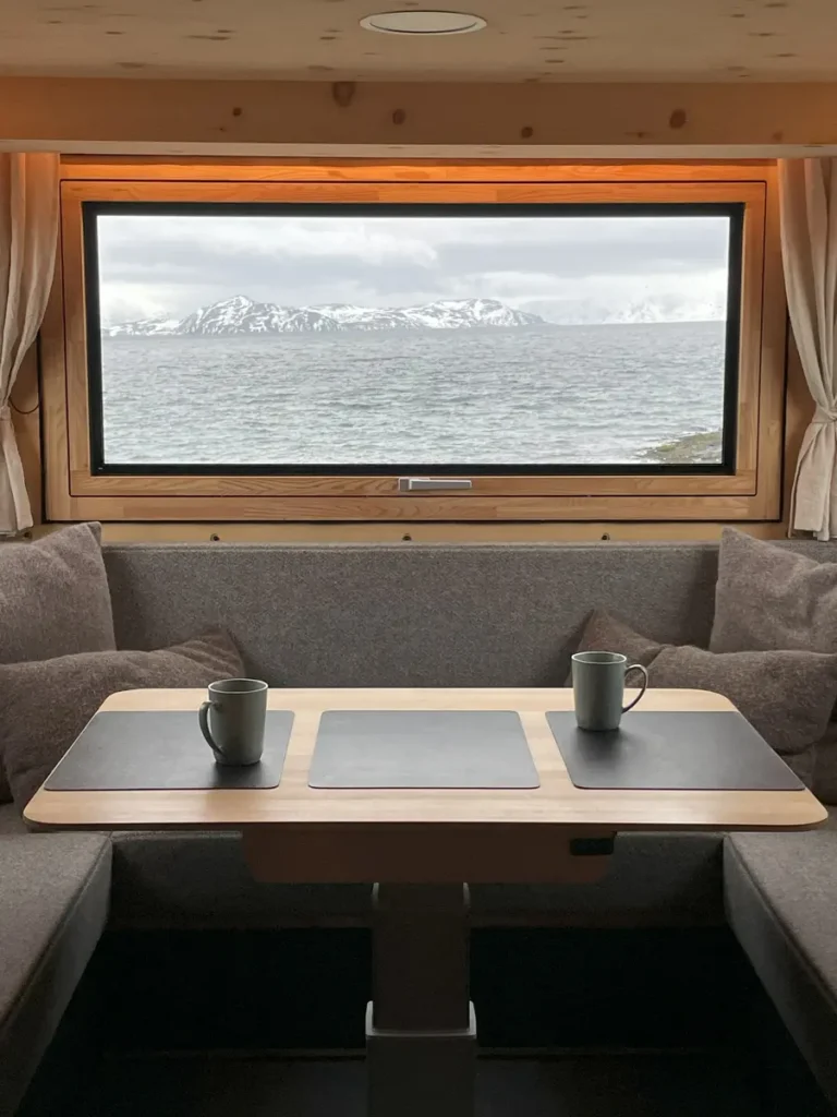 Selbstausbau-Interieur mit Holzwänden, Tisch, Sitzbank und Panoramafenster mit Blick auf Meer und Gebirge.