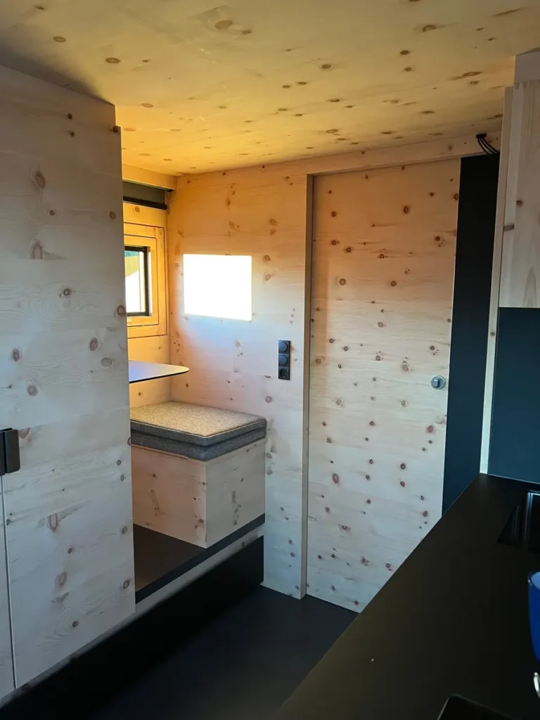 Innenansicht eines minimalistischen Campervans mit hellem Holzausbau, Sitzbank und Küchenzeile.