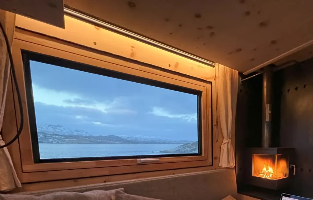 Gemütlicher Campervan-Selbstausbau mit Holzofen und Blick auf winterliche Fjordlandschaft.