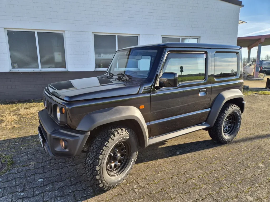 Schwarzer Suzuki Jimny 4x4 mit Offroad-Reifen parkt vor einem Gebäude.