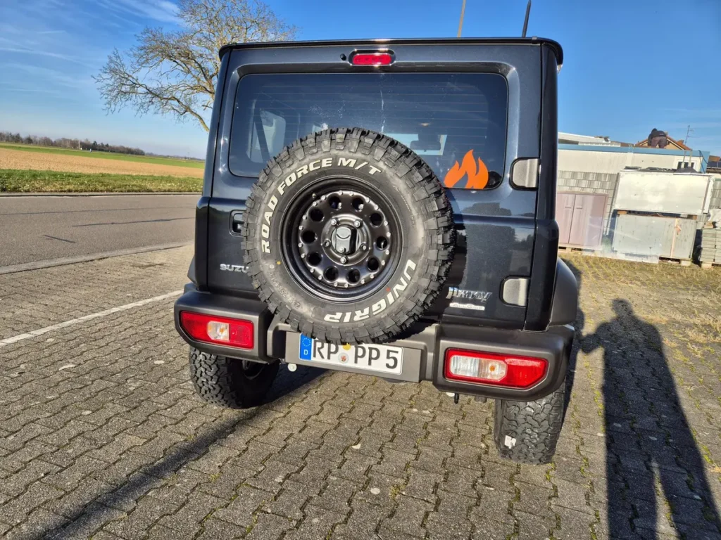Rückansicht eines grauen Suzuki Jimny 4x4 mit Reserverad und Offroad-Bereifung.