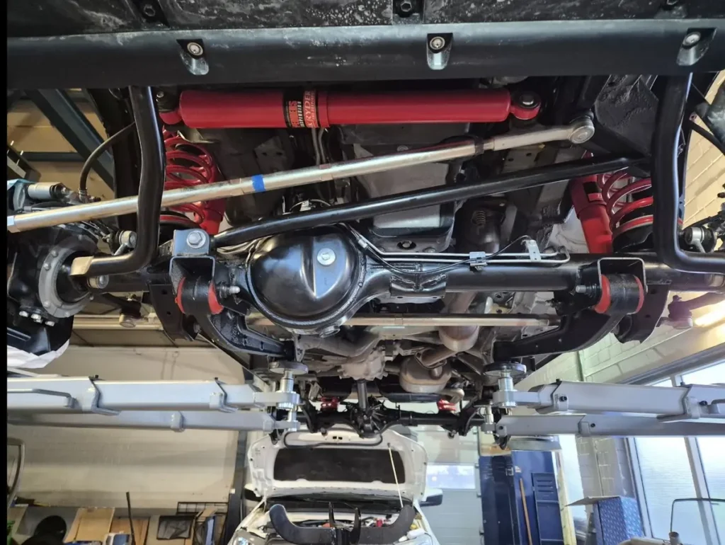 Unterseite eines 4x4 Geländewagens mit rotem Offroad-Fahrwerk, Stoßdämpfern und verstärkter Hinterachse.