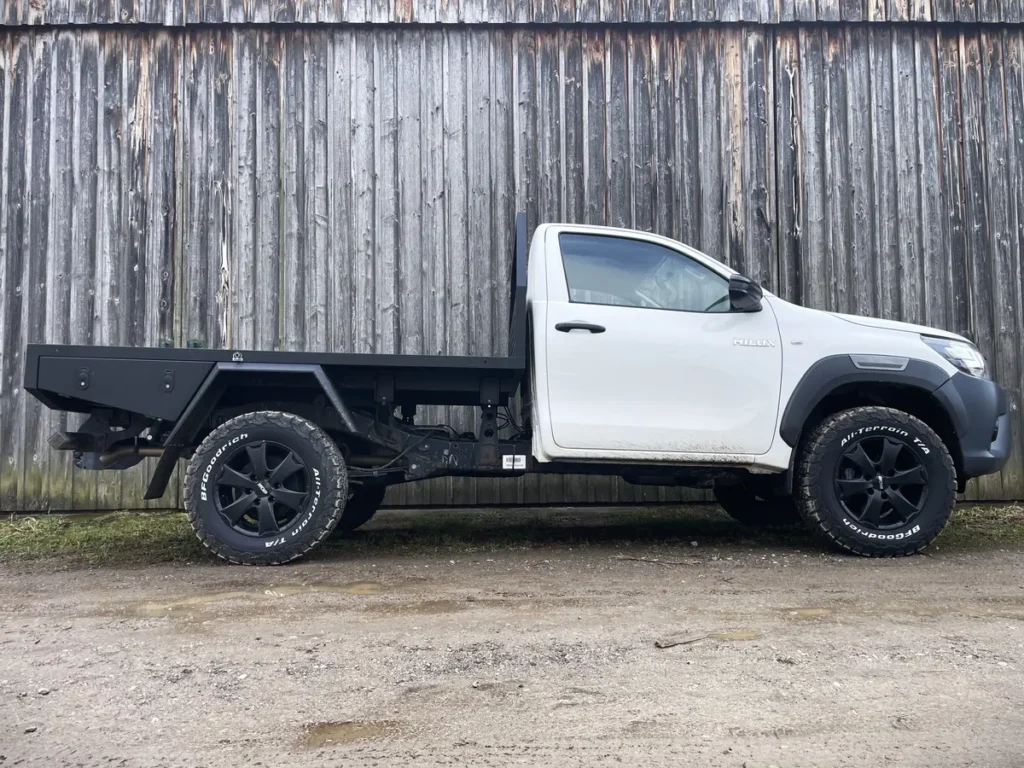 Ein weißer Toyota Hilux Pick-up 4x4 mit schwarzer Expeditionspritsche vor einer Holzwand.