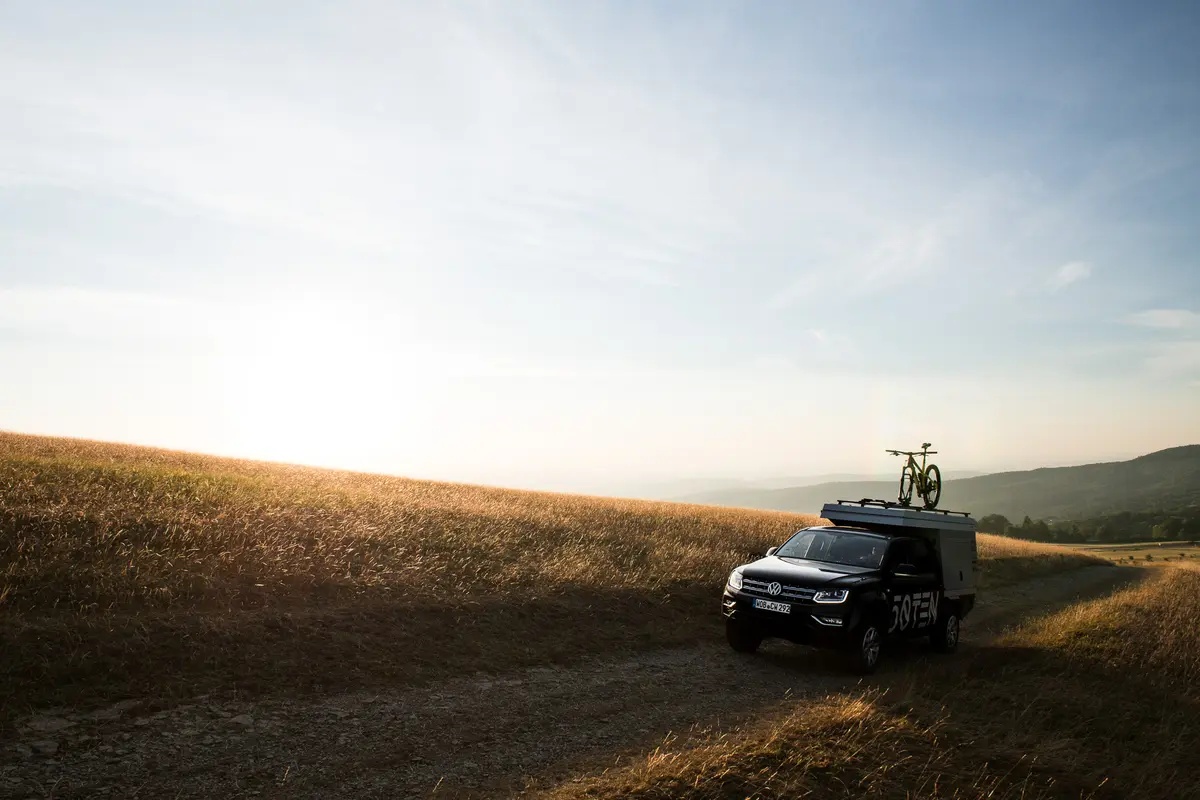 Ein schwarzer VW Amarok 4x4 Pickup mit Wohnkabine und Mountainbike fährt auf einem Feldweg.
