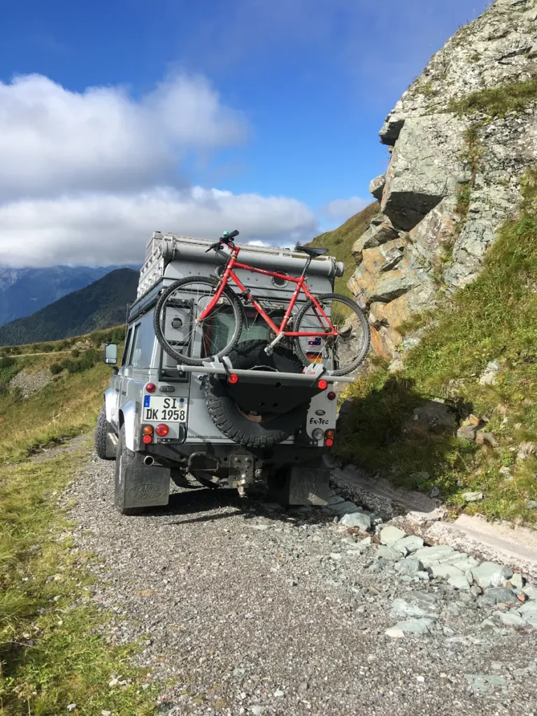 Land Rover Defender 4x4 mit Fahrrad auf Heckträger in alpiner Schotterlandschaft.