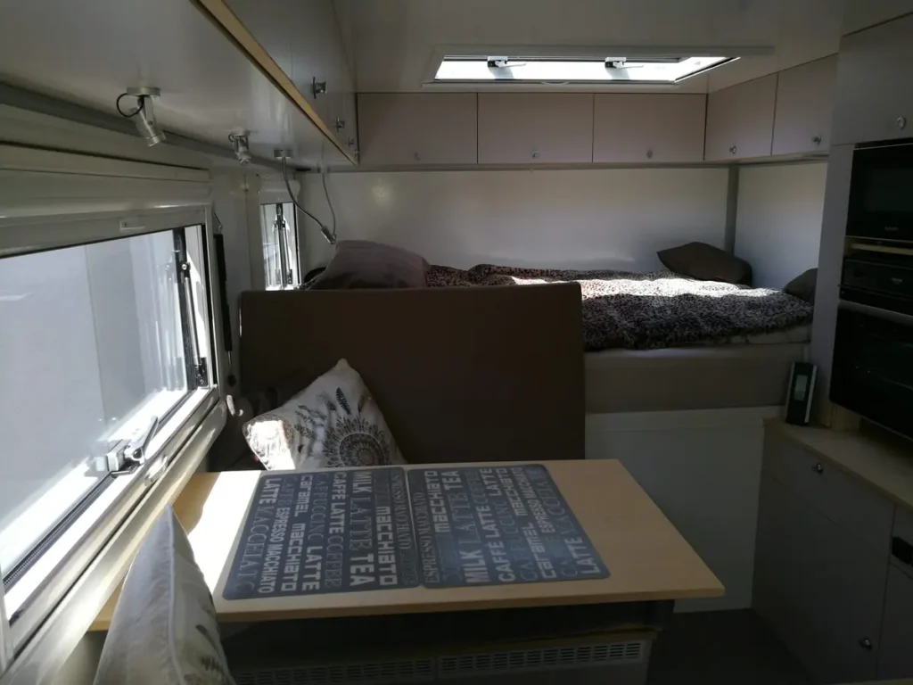 Heller Innenraum eines Campervans mit Bett, Tisch und integrierter Küchenzeile, im Stil eines Selbstausbaus.