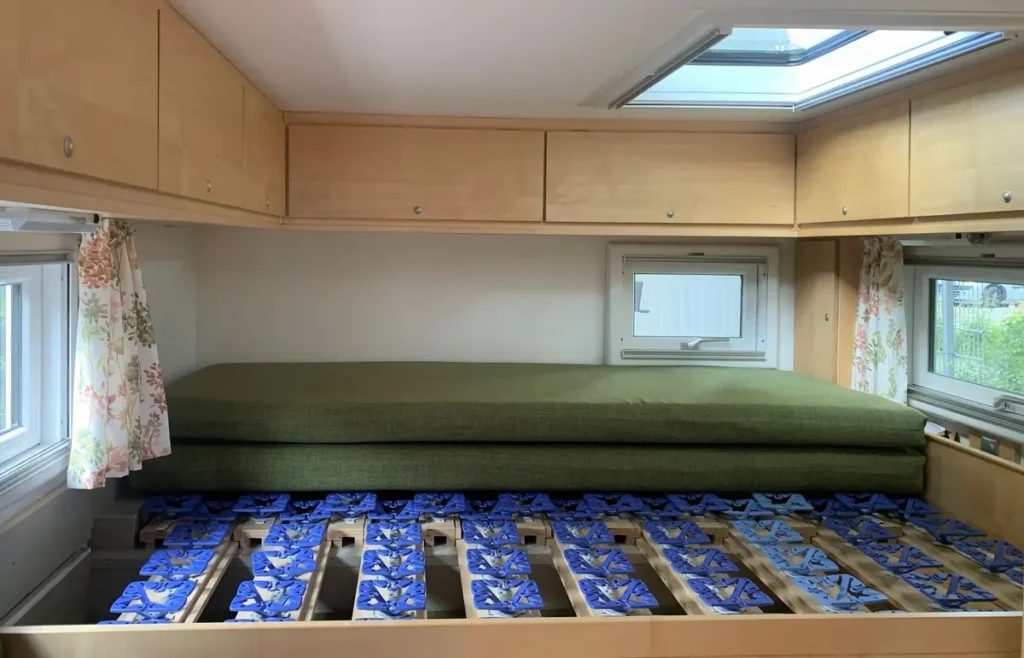 Innenansicht eines Campervan-Betts mit verstellbarem Lattenrost, gestapelten Matratzen, Hängeschränken und Fenstern.