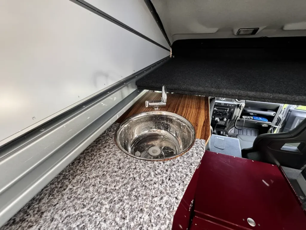 Kompakte Küchenzeile in einem 4x4-Selbstausbau mit Edelstahlspüle und roter Kühlbox.