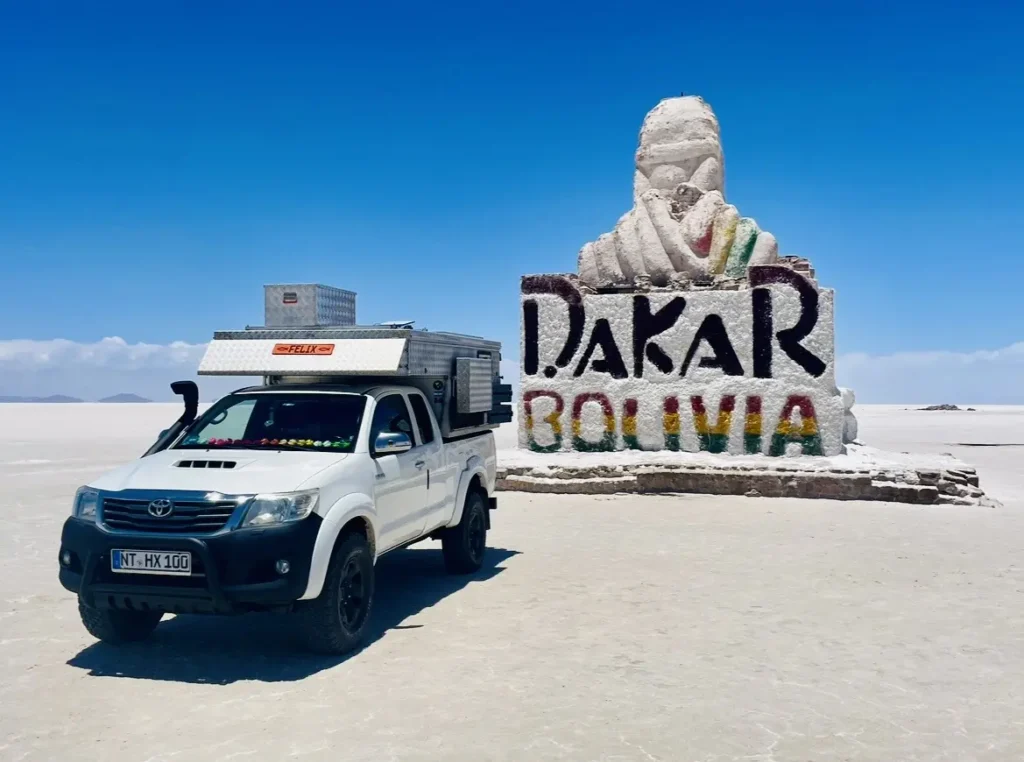 Ein weißer 4x4 Pick-up mit Wohnkabine und Schnorchel auf einem Salzsee vor dem Dakar-Monument.