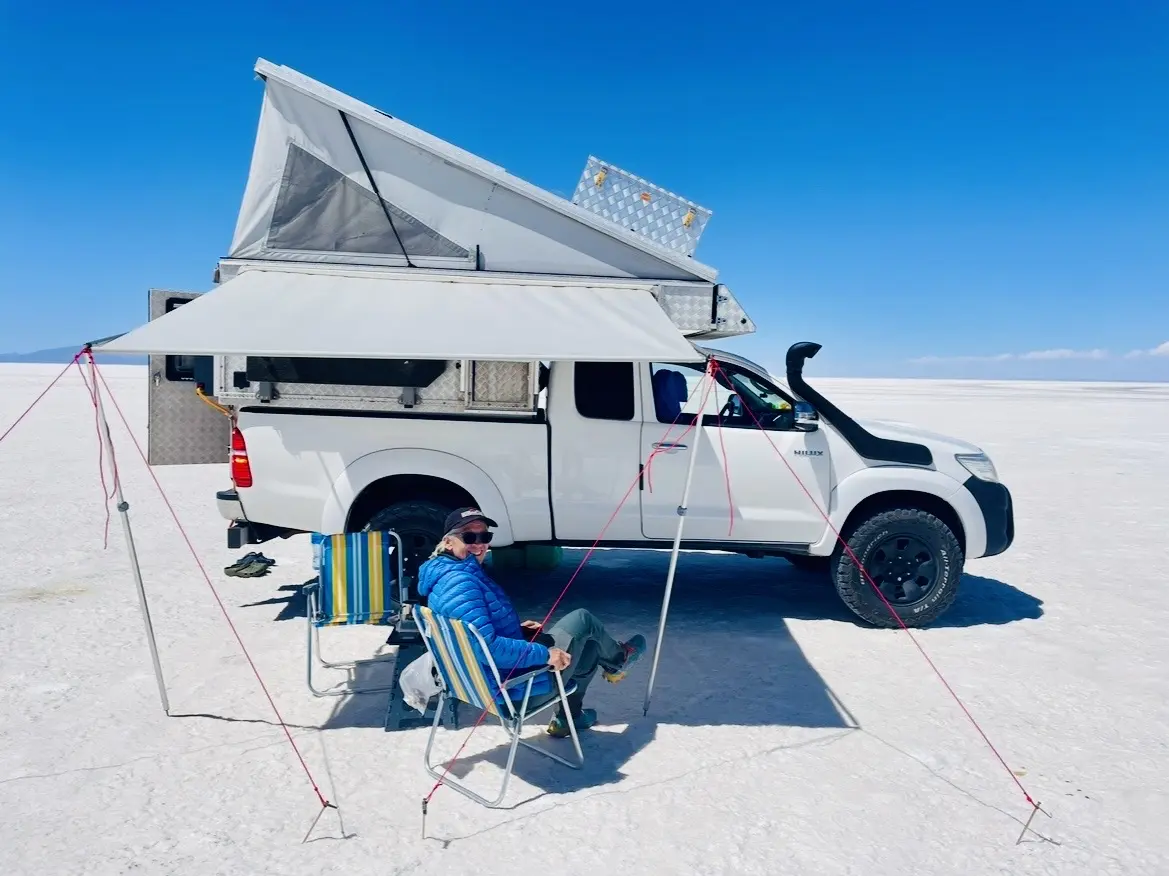 Ein 4x4 Pick-up mit Wohnkabine und Markise steht auf einer Salzfläche, davor sitzt eine Person.