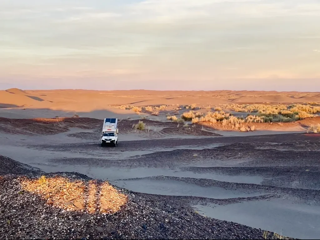 Pick-up Wohnkabine mit Solarmodulen fährt durch eine sandige Wüstenlandschaft bei Sonnenuntergang.