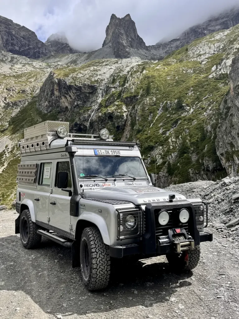 Expeditionsfahrzeug Land Rover Defender auf Schotterpiste vor beeindruckender Bergkulisse mit Wasserfällen.