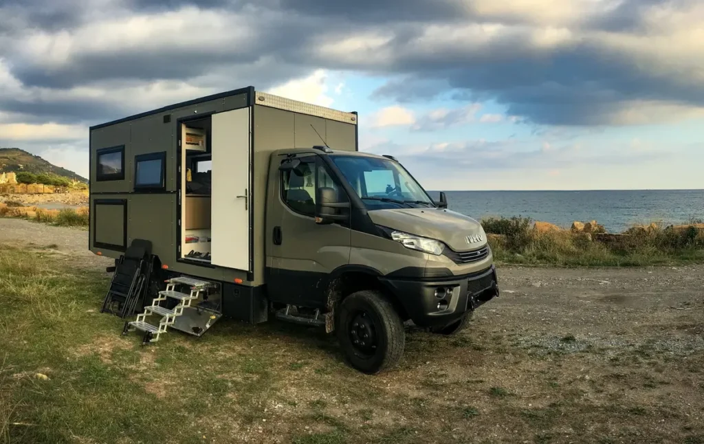 Ein olivgrünes Iveco 4x4 Expeditionsmobil am Meer mit offener Tür und ausgefahrenen Stufen.
