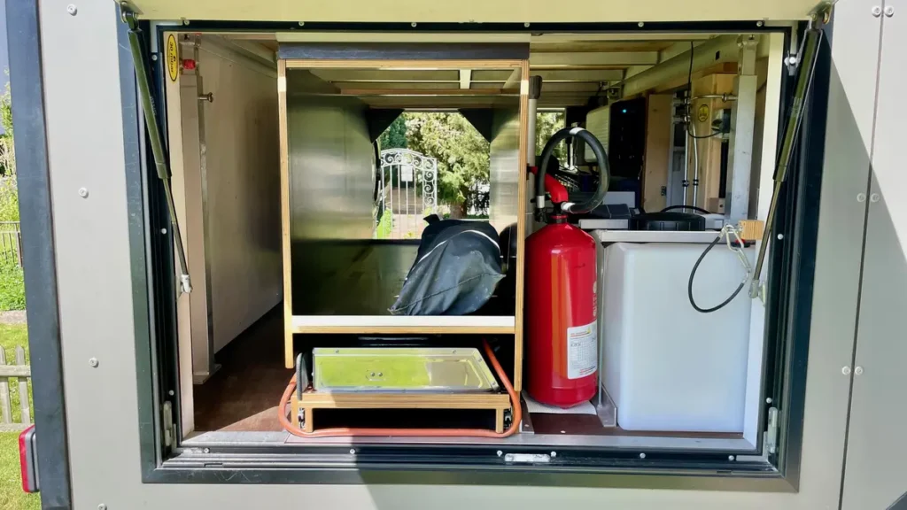 Heckbereich eines Campingtrailers mit integrierter Outdoor-Küche, Frischwassertank und Feuerlöscher.