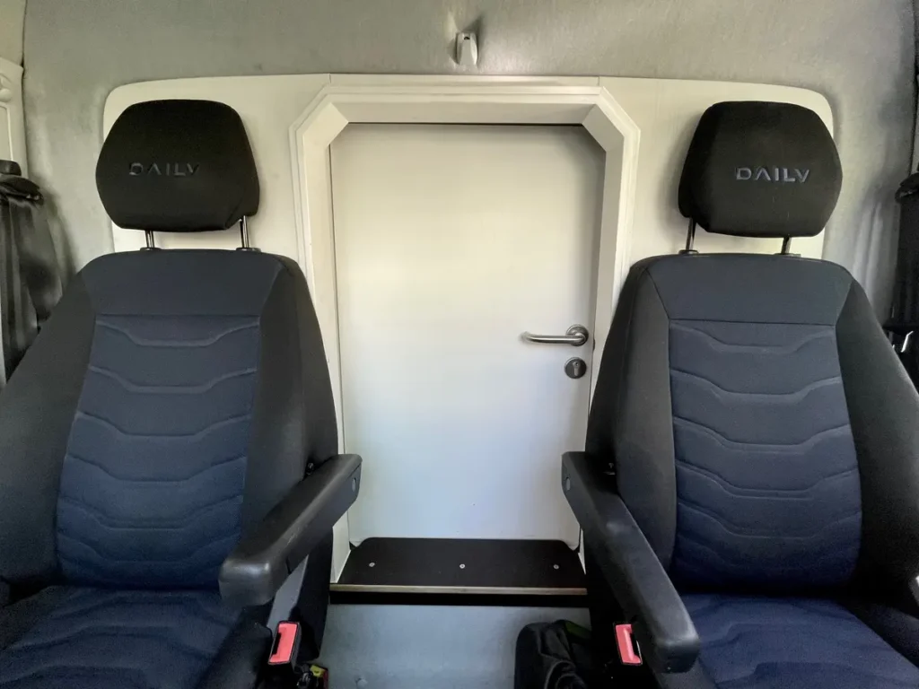 Innenansicht eines Selbstausbau-Campervans mit zwei Iveco Daily Sitzen und einer Verbindungstür.