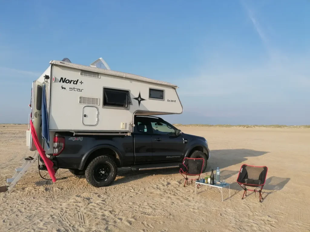 Dunkelgrauer Pick-up mit weißer Wohnkabine steht auf sandigem Strand, daneben Campingtisch und Stühle.