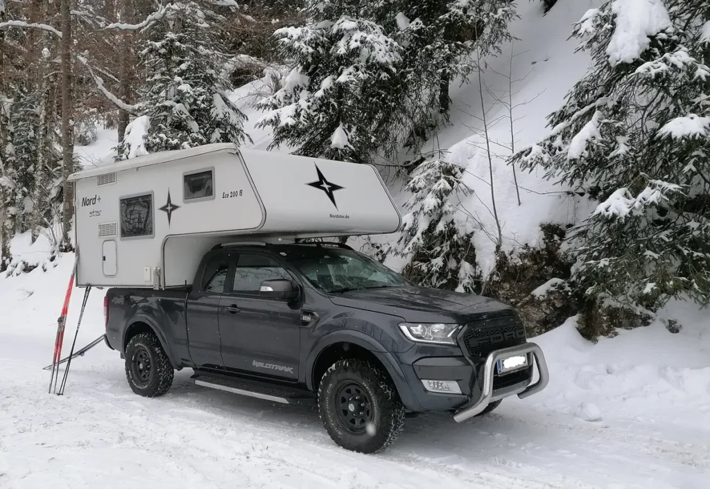 Ein grauer 4x4 Pick-up mit Wohnkabine und Skiern in verschneiter Winterlandschaft.