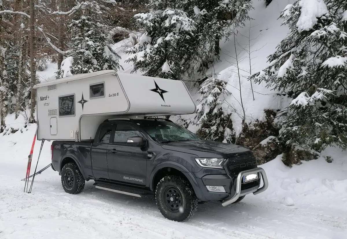 Ein grauer 4x4 Pick-up mit Wohnkabine und Skiern in verschneiter Winterlandschaft.