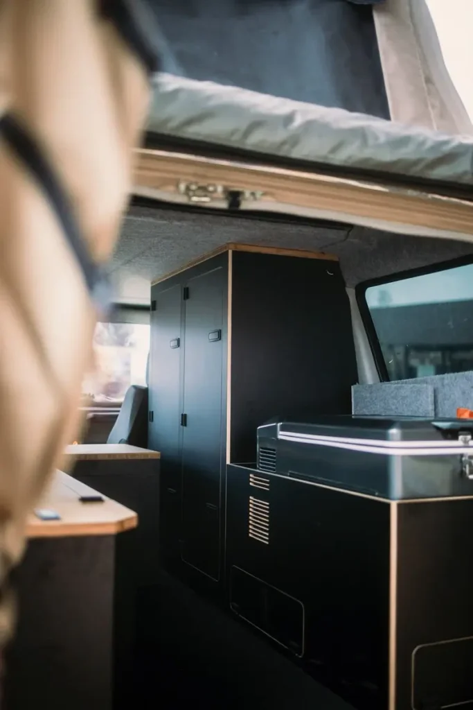 Detailansicht des Innenausbaus eines Campervans mit dunklen Möbeln, integrierter Kühlbox und Aufstelldach.
