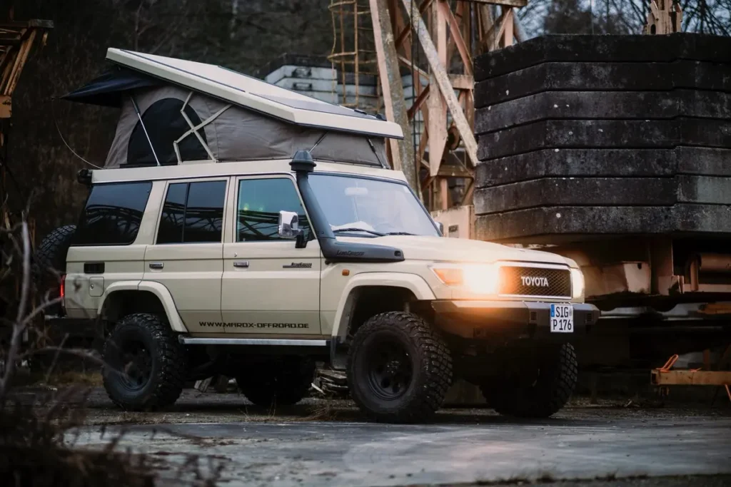 Ein helles Expeditionsmobil mit Pop-Up-Dachzelt und Schnorchel steht in einer industriellen Offroad-Umgebung.