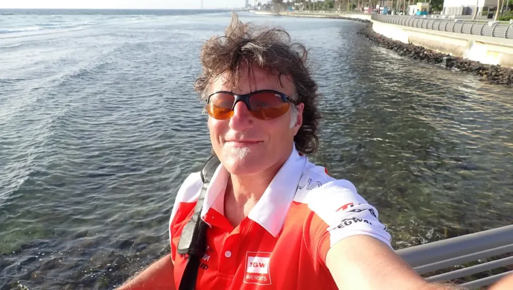 Ein lächelnder Mann mit Sonnenbrille am Wasser, mit Küstenlandschaft im Hintergrund.