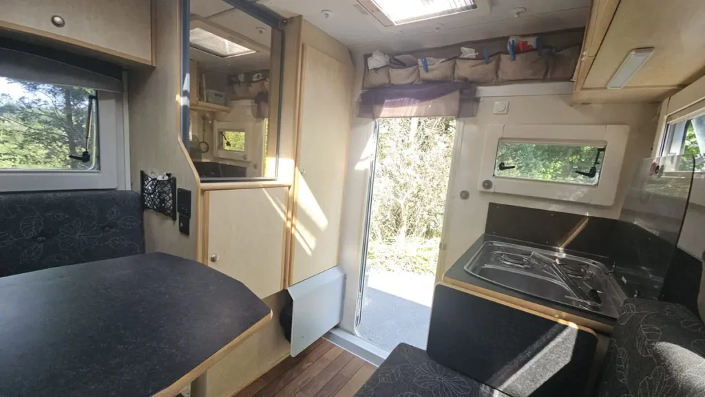 Innenansicht eines Campervans mit integrierter Küche, Esstisch, Sitzbereich und offener Tür nach draußen.