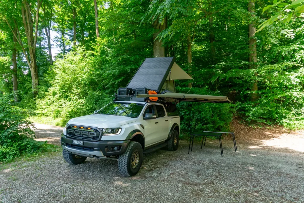 Ein weißer Ford Ranger Pick-up mit Dachzelt, Solarmodul und Markise im Wald geparkt.