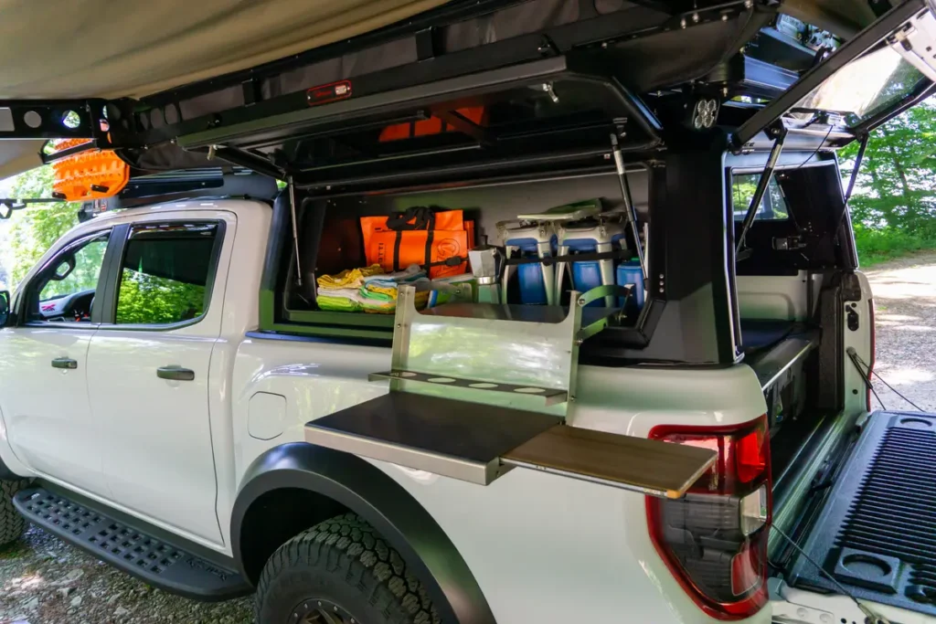 Weißer Pick-up mit offener Pick-up Wohnkabine, Dachzelt und ausziehbarer Campingküche im Gelände.
