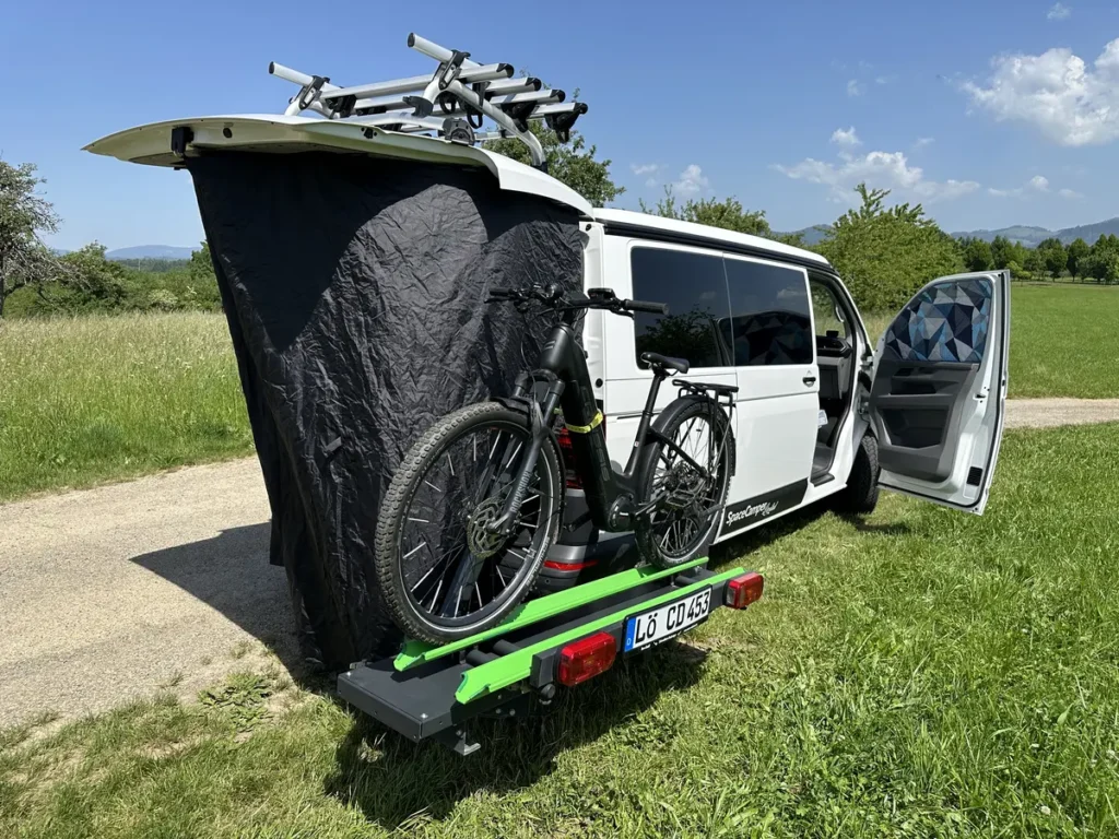 Ein weißer Campervan mit Heckanbauzelt und zwei Fahrrädern auf einem Heckträger.