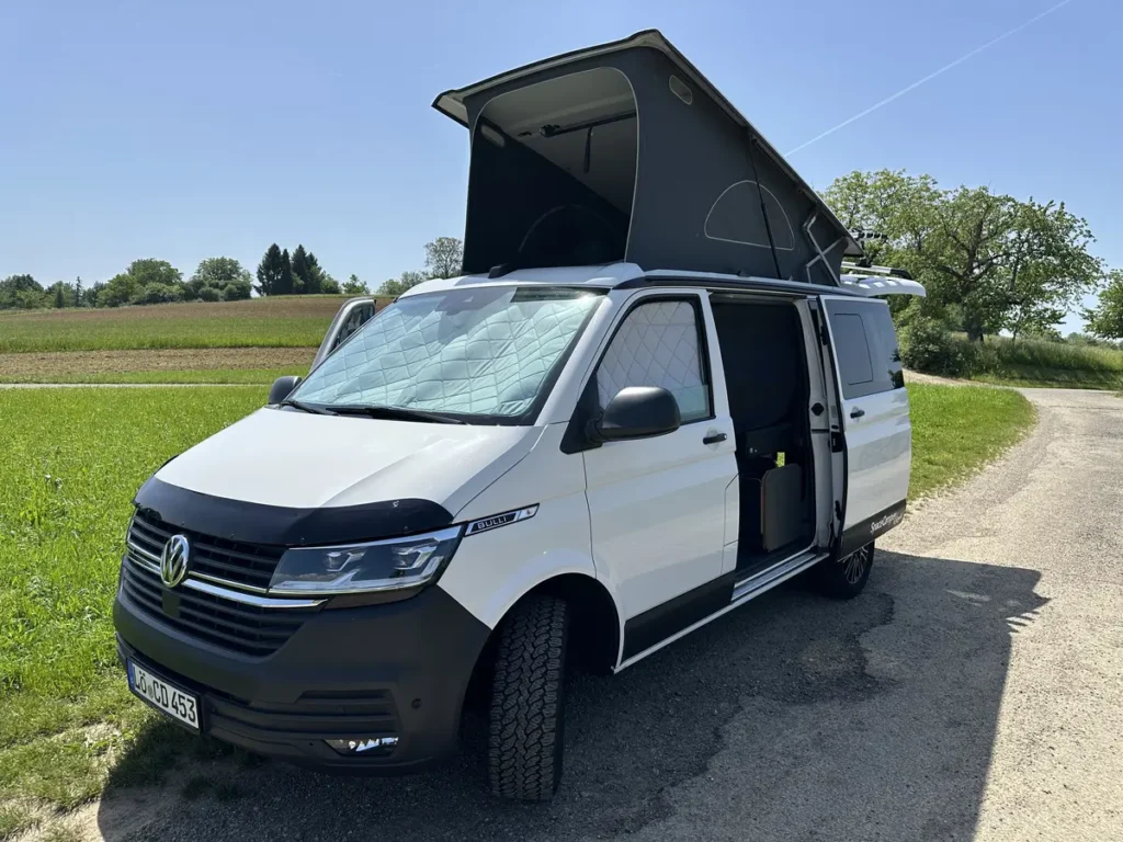 Ein weißer Campervan mit offenem Aufstelldach und Offroad-Reifen steht auf einem Feldweg.