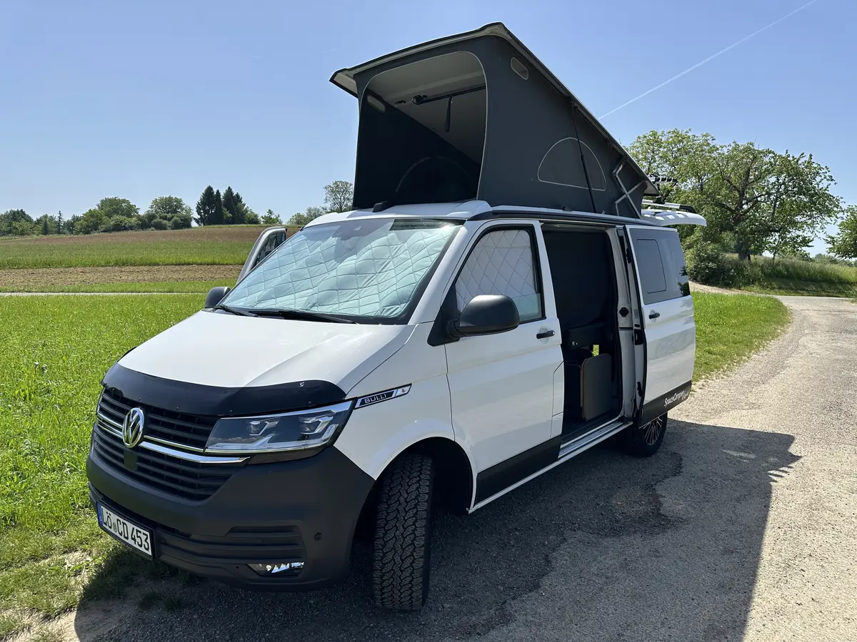 Ein weißer Campervan mit offenem Aufstelldach und Offroad-Reifen steht auf einem Feldweg.