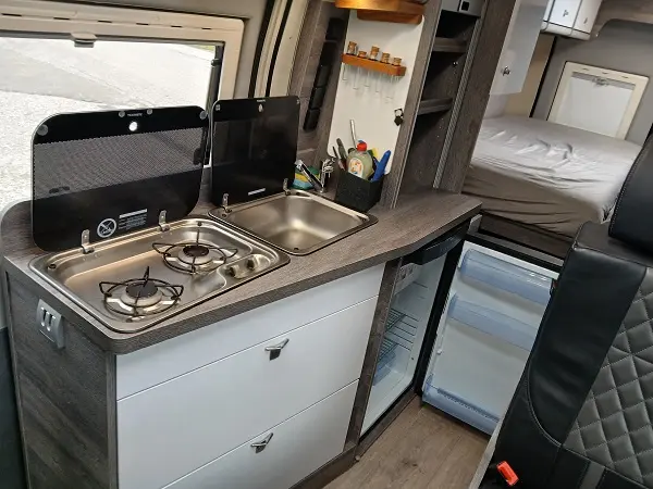 Innenansicht eines Selbstausbau-Vans mit voll ausgestatteter Küchenzeile und Bett.