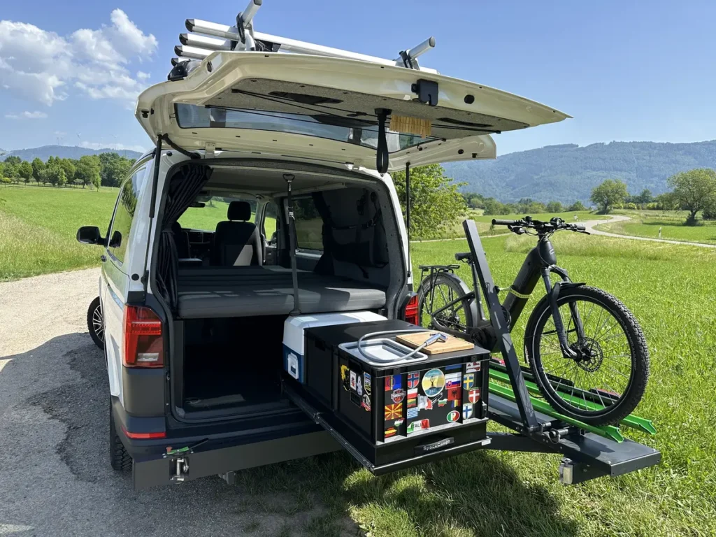 Weißer Campervan mit offenem Heck, Heckauszug, Fahrradträger und E-Bike in grüner Landschaft.