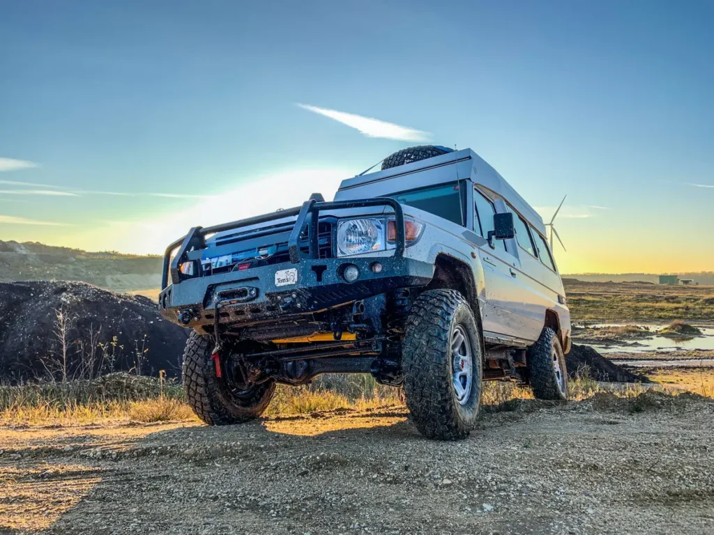 Weißer Toyota Land Cruiser 4x4 Campervan mit Offroad-Bereifung und Bullbar in staubiger Landschaft.