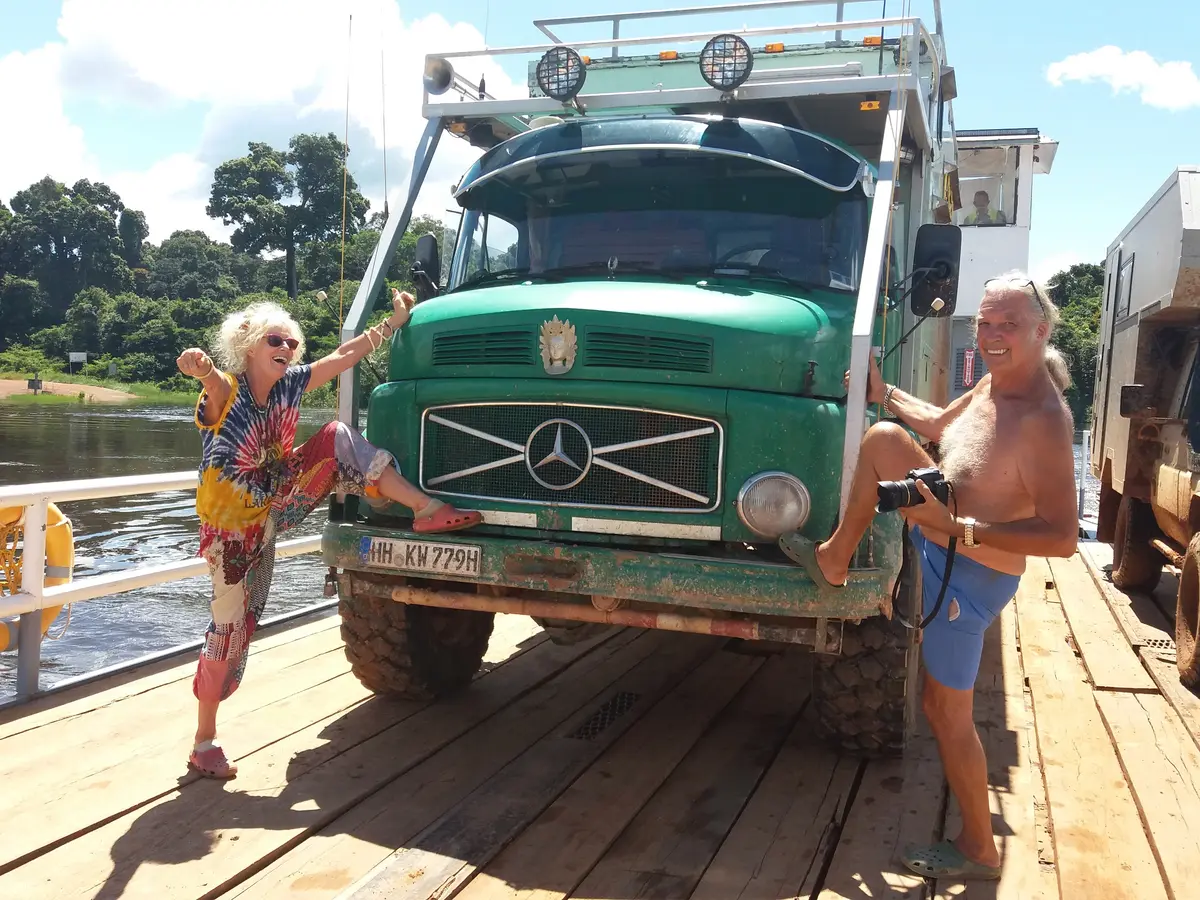 Fröhliche Overlander posieren auf einer Flussfähre vor ihrem grünen Mercedes 4x4 Expeditionsmobil.