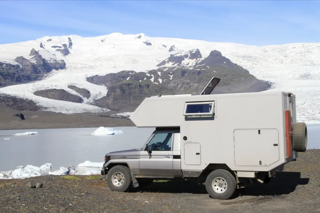 Graues Allrad Expeditionsmobil mit Wohnkabine parkt an einem Gletschersee mit Eisbergen und schneebedeckten Bergen.
