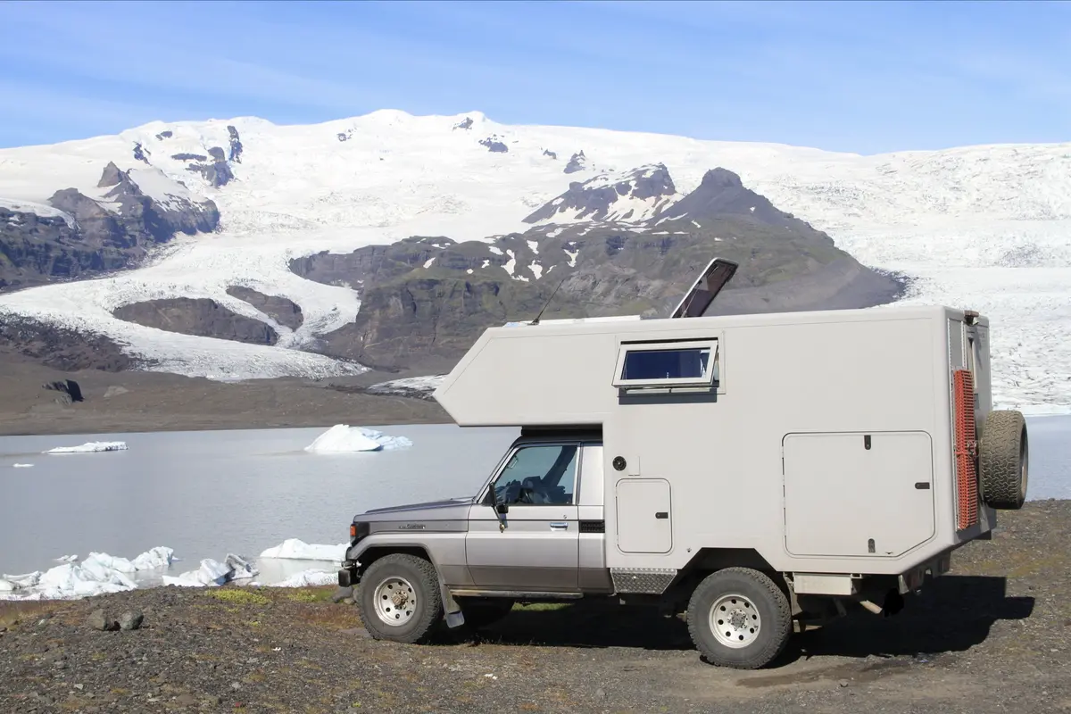 Graues Allrad Expeditionsmobil mit Wohnkabine parkt an einem Gletschersee mit Eisbergen und schneebedeckten Bergen.