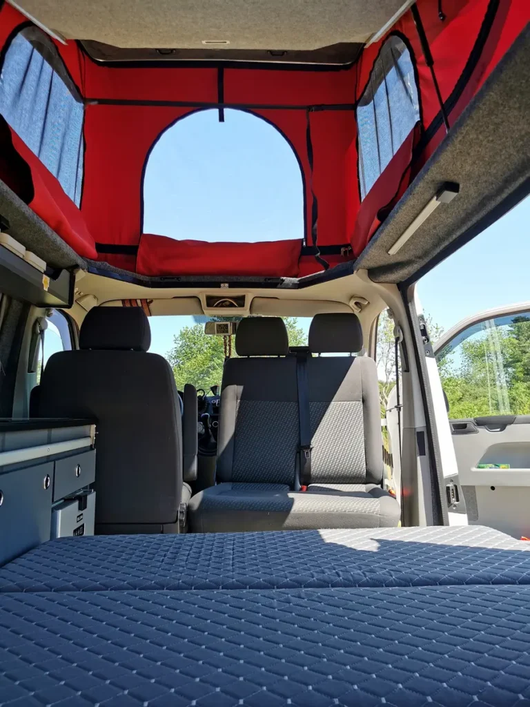 Innenansicht eines Campervans mit ausgefahrenem Aufstelldach, gemütlichem Bett und Küchenmodul.