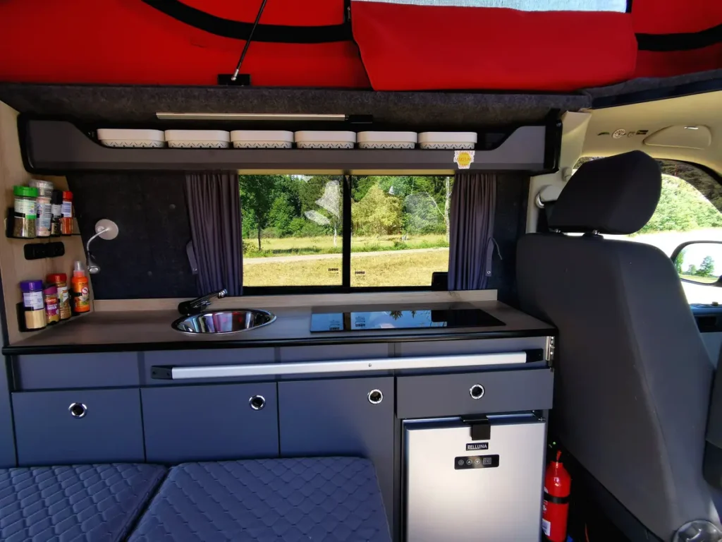 Innenansicht eines maßgeschneiderten Campervan-Ausbaus mit Küchenblock, Spüle, Kochfeld und Kompressorkühlschrank.