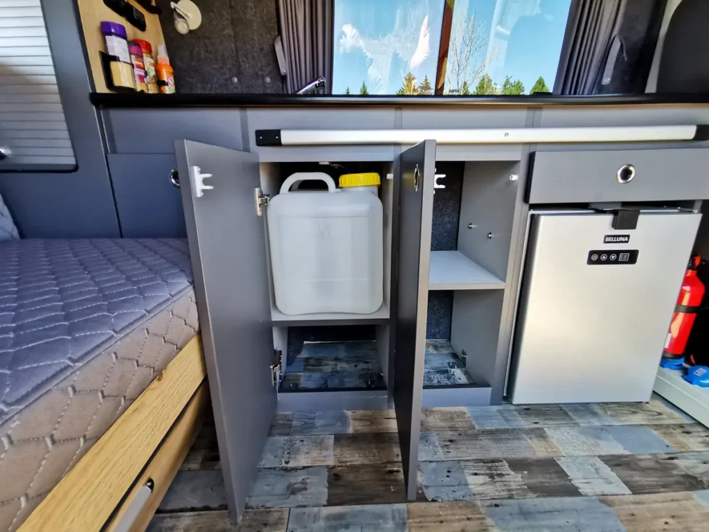 Innenansicht eines Campervans mit Küchenzeile, integriertem Kühlschrank, Wassertank und Schlaffläche.