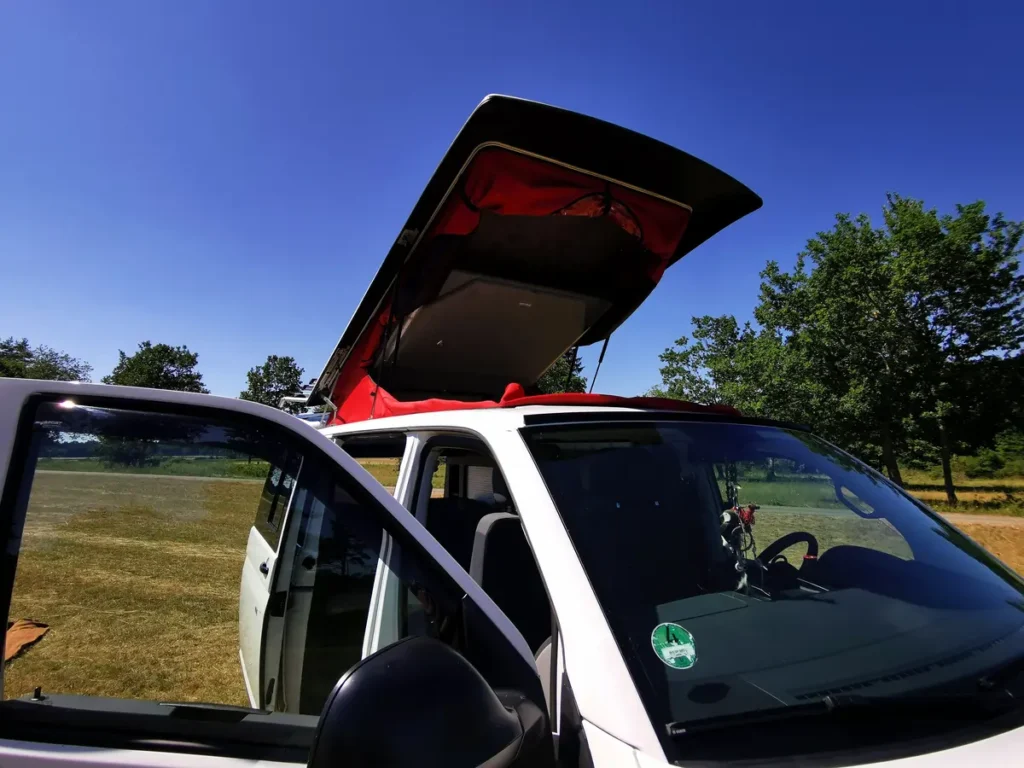 Weißer Campervan mit ausgefahrenem Aufstelldach und offenen Türen auf einer Wiese.