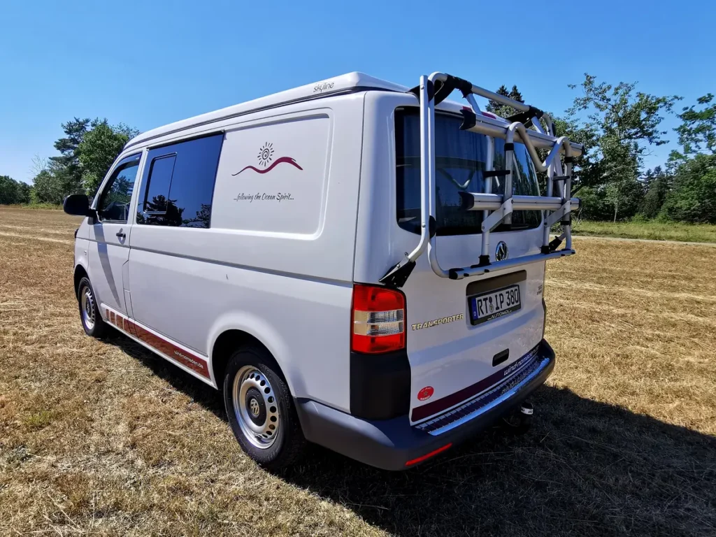 Weißer VW Campervan mit Aufstelldach und Fahrradträger auf einem trockenen Feld.