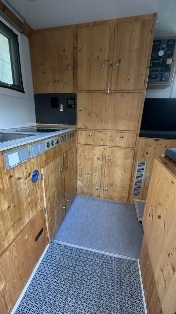 Holzverkleidetes Interieur eines Campervans mit Küche, Staufächern und Elektrik-Panel.