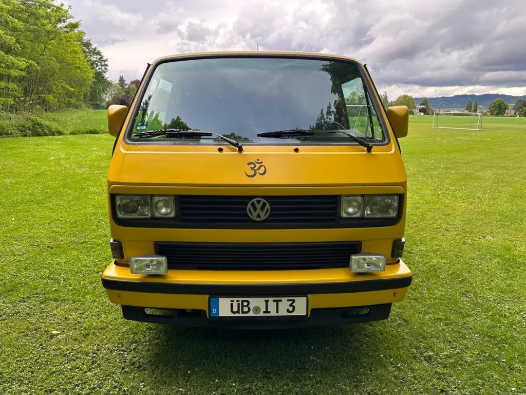 Ein gelber VW T3 Campervan steht frontal auf einer grünen Wiese unter bewölktem Himmel.