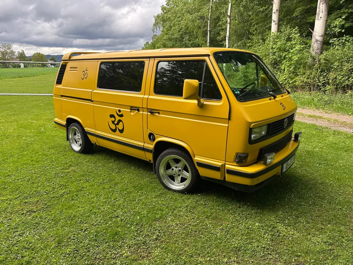 Klassischer gelber VW T3 Bulli als Selbstausbau auf einer grünen Wiese am Waldrand.
