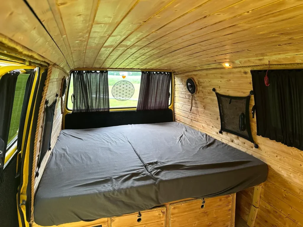 Gemütlicher Innenraum eines Campervans mit Holzverkleidung, großem Bett und praktischem Stauraum.