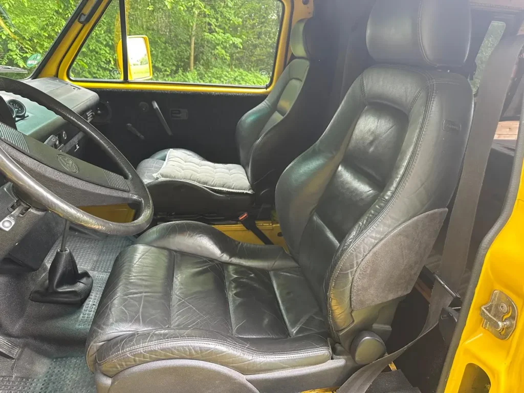 Innenraum eines gelben Campervans mit schwarzen Ledersportsitzen, Schalthebel und VW-Lenkrad.