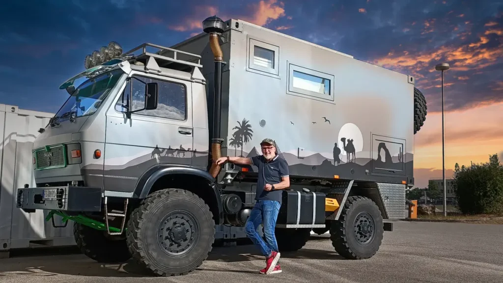 Ein lächelnder Mann steht neben seinem grauen MAN 4x4 Expeditionsmobil mit Wüstendekor.